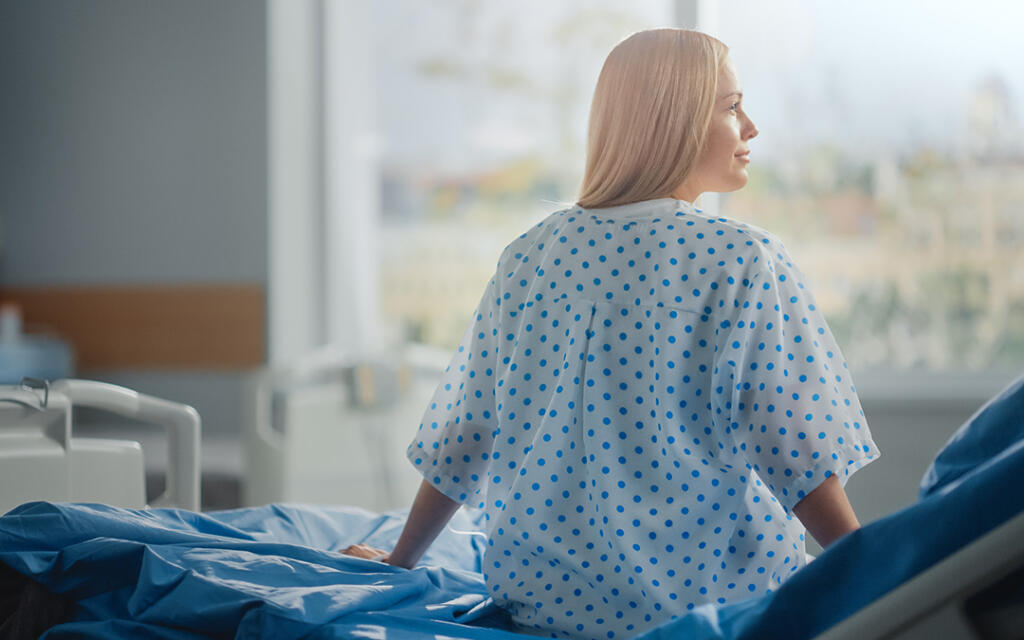 Patient in surgical gown sitting on edge of bed looking out of a window
